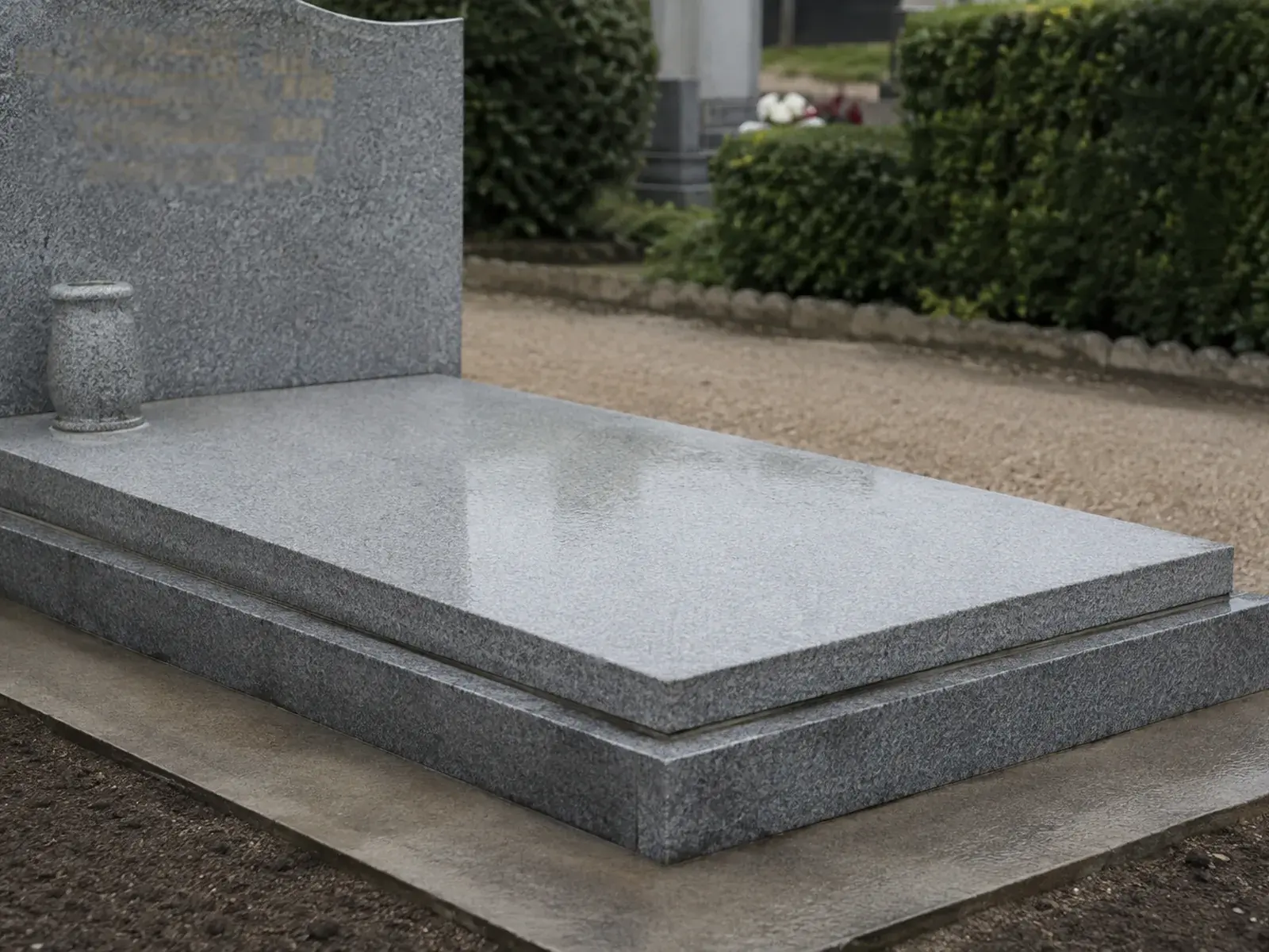 Sépulture en granit gris clair fraîchement nettoyée, pierre encore humide du rinçage, abords dégagés le long d’une allée de gravier