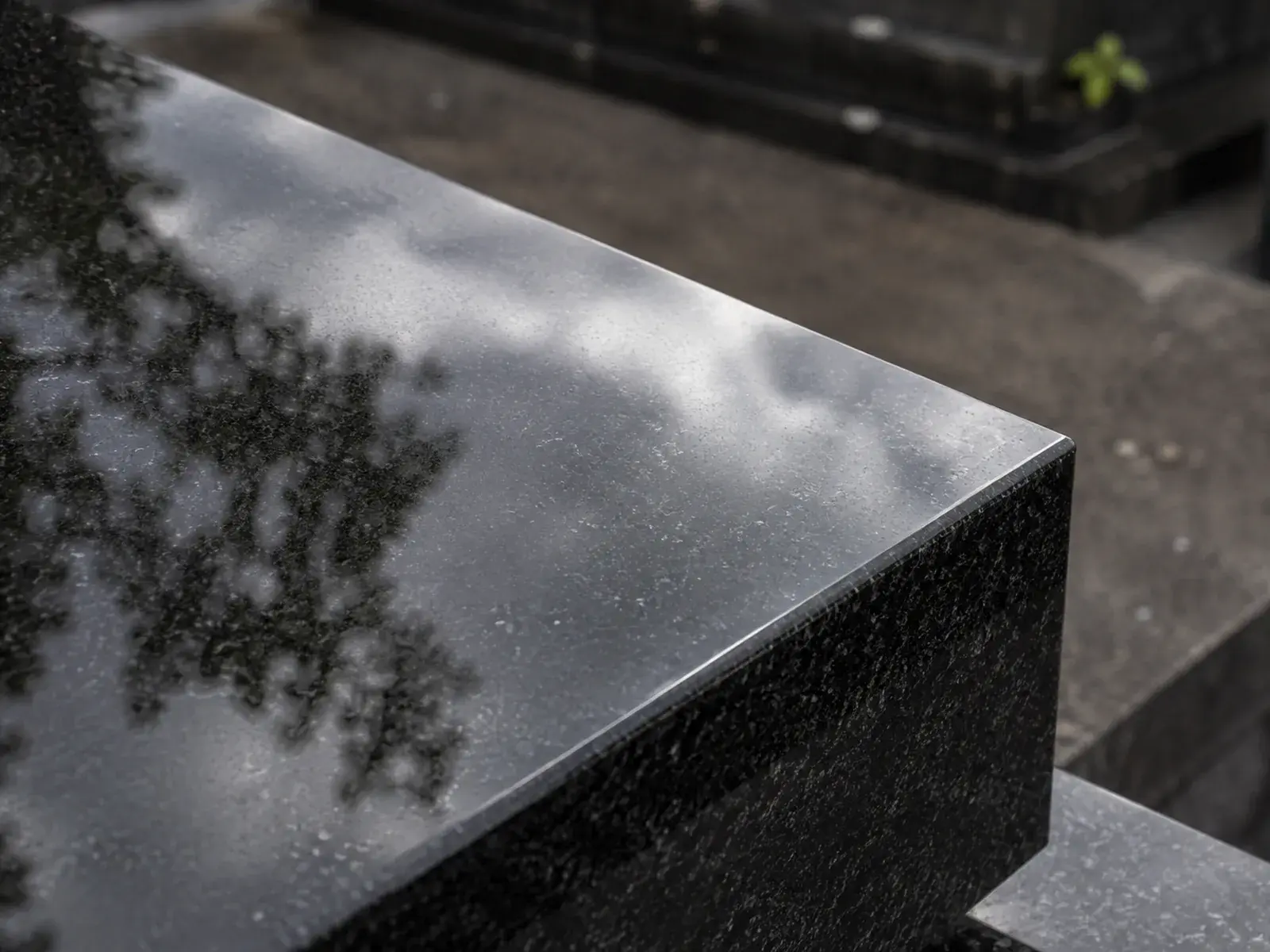 Arête d’un monument en granit noir poli, dont la surface reflète un ciel nuageux et la silhouette d’un arbre